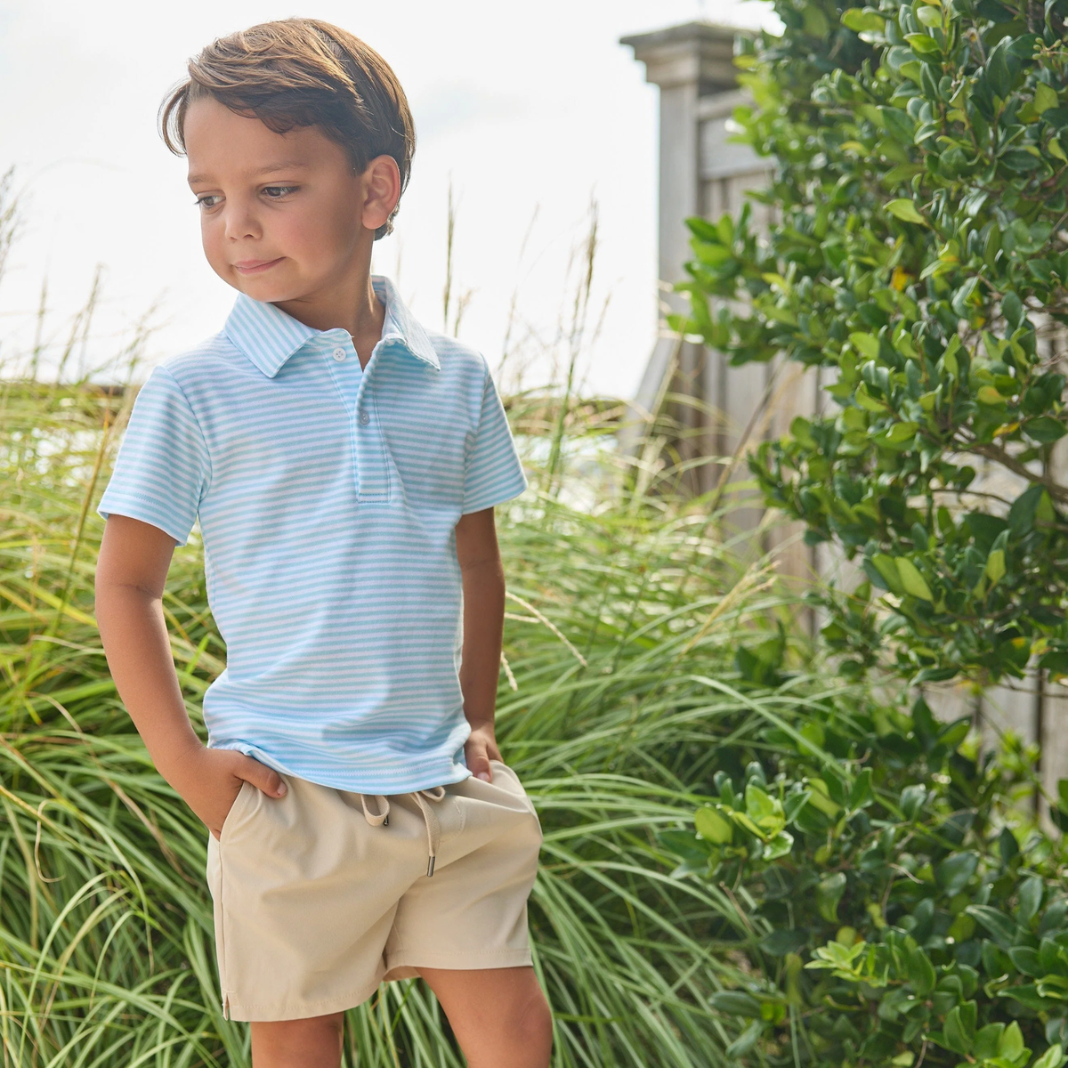 Young boy wearing a light blue striped polo shirt and beige shorts standing outdoors with greenery around.
