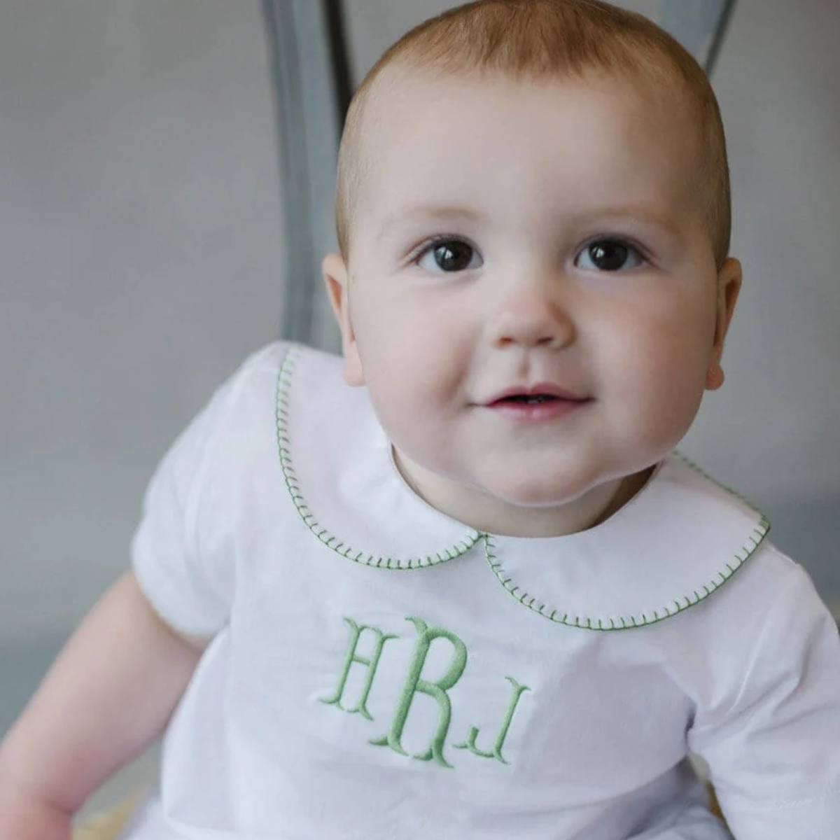 Baby wearing a white outfit with green collar trim and monogram, sitting against a neutral background.