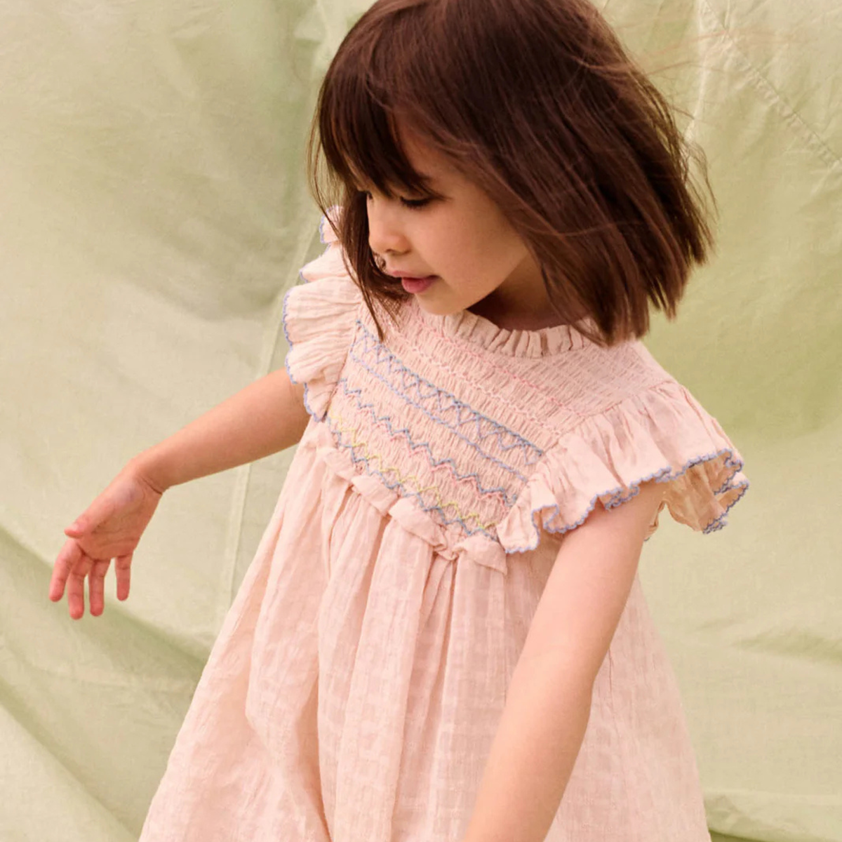 Young girl wearing a pink dress with an embroidered bodice and ruffle sleeves on a green background. 
