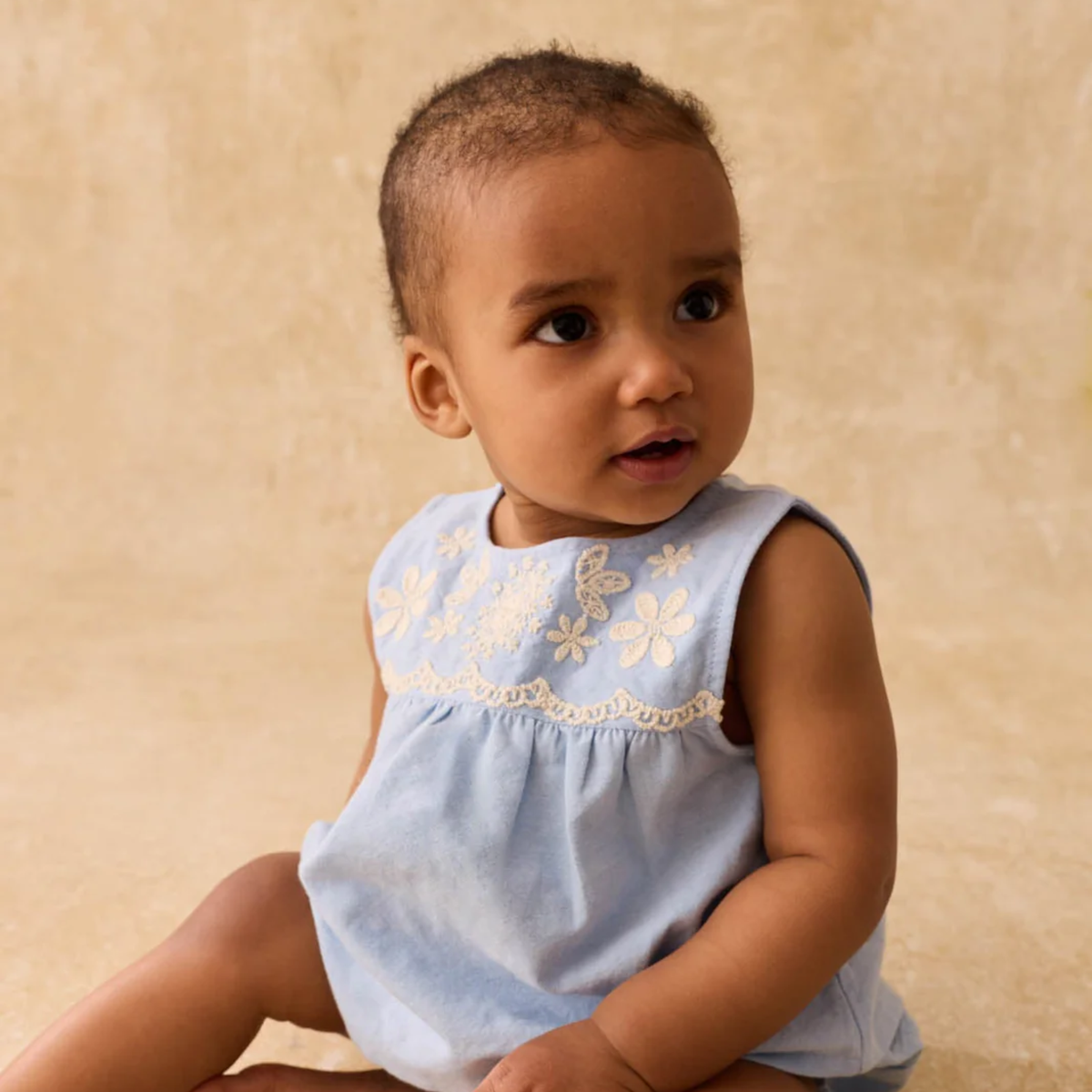 Baby wearing a light blue blouse with floral embroidery on a beige background.