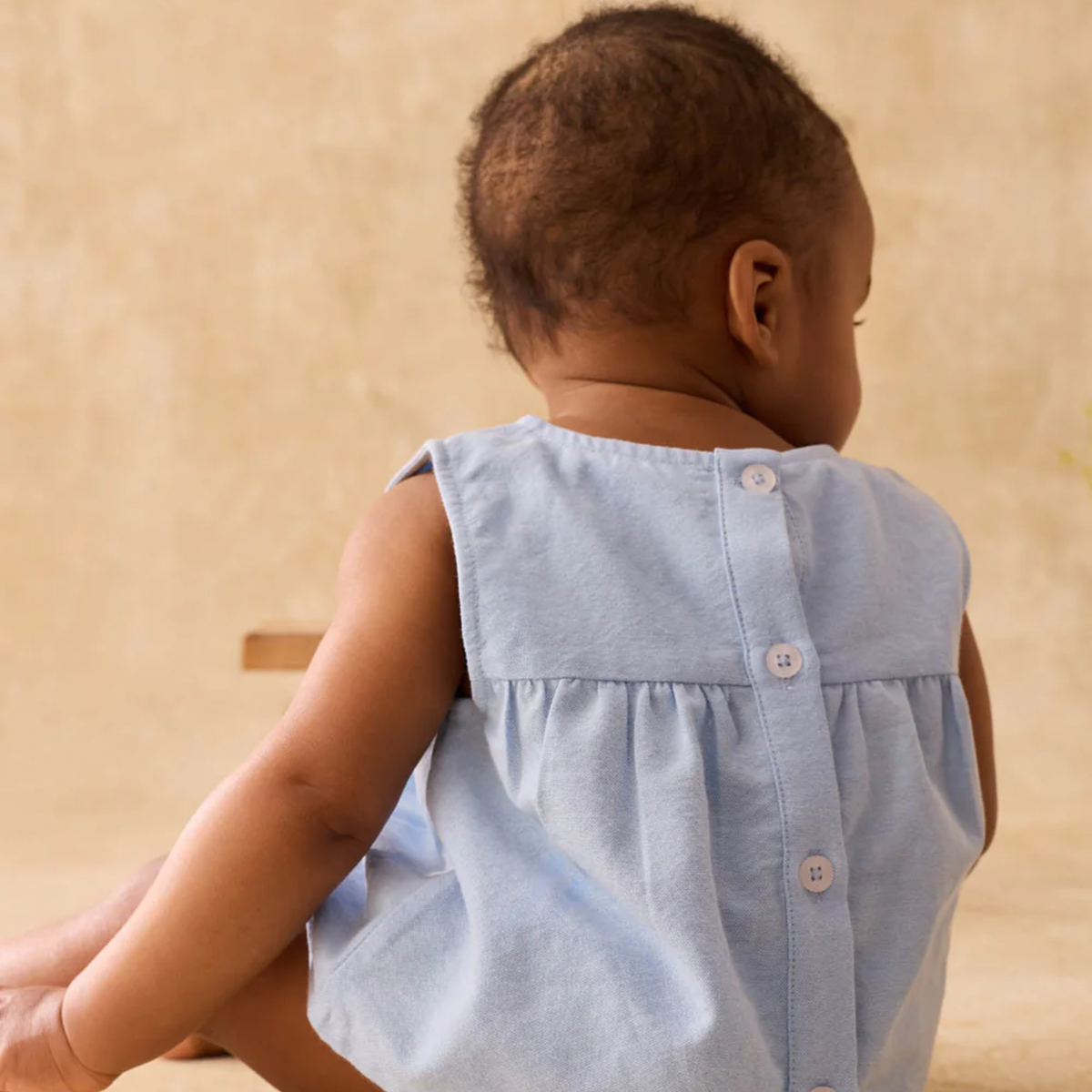 Child wearing a light blue sleeveless blouse with back buttons on a beige background.