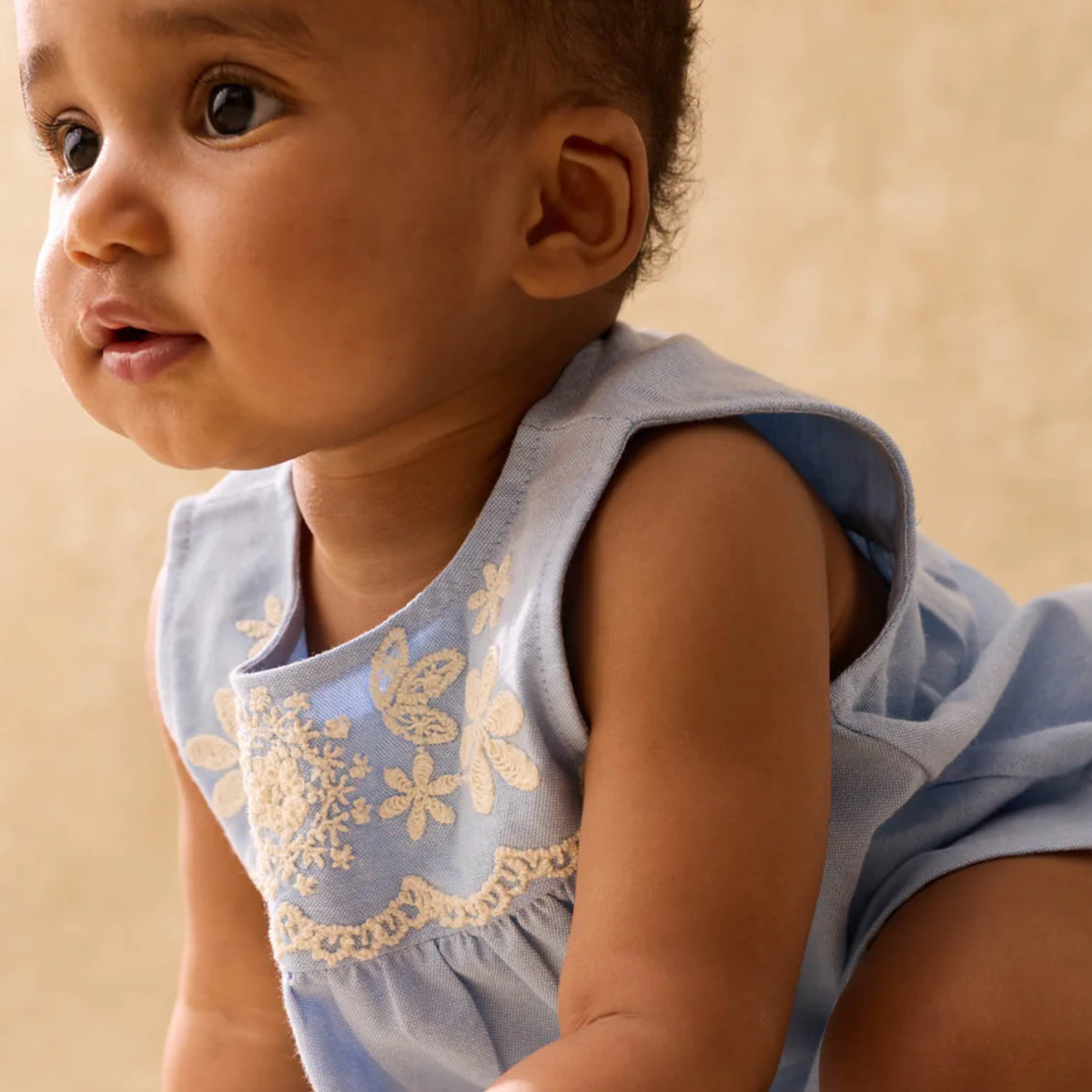 Baby wearing a light blue blouse with floral embroidery on a beige background.
