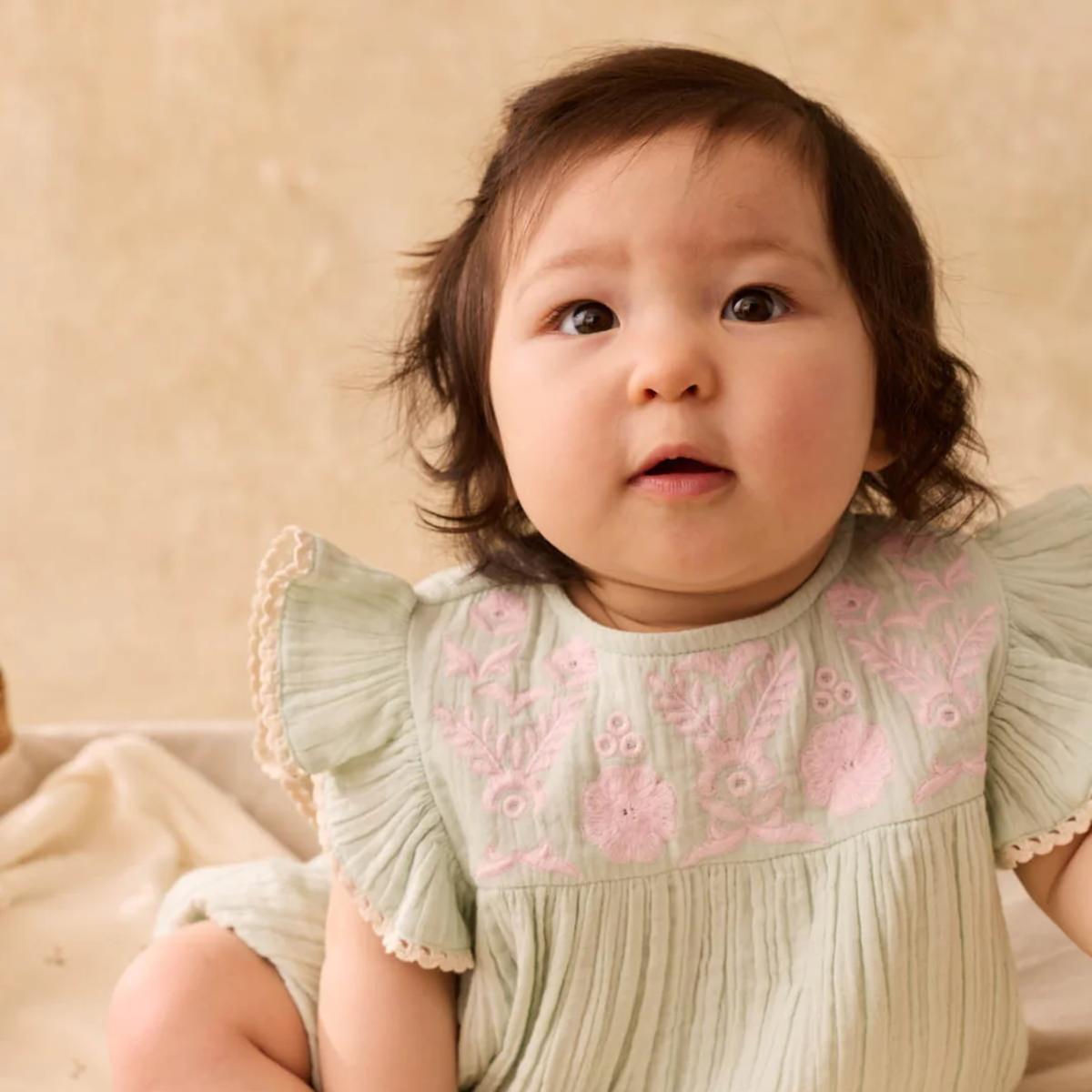 Baby wearing a light green romper with pink floral embroidery against a beige background.