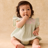 Baby in a light green romper with pink floral embroidery on a beige background.