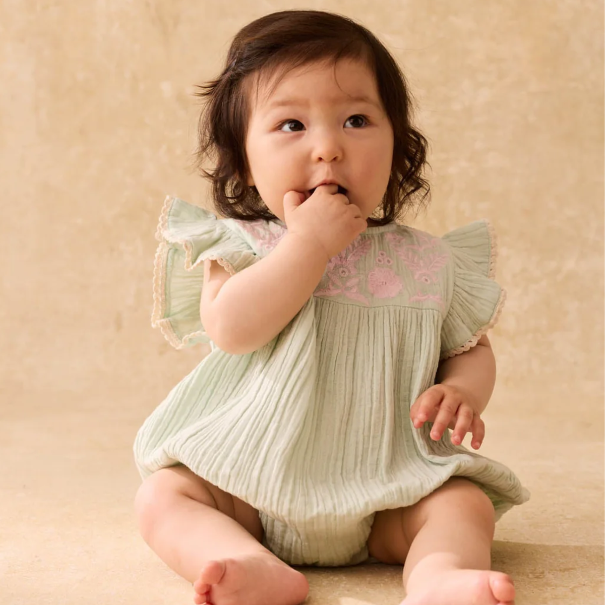 Baby in a light green romper with pink floral embroidery on a beige background.