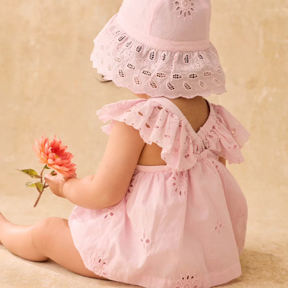 Baby in a pink lace outfit including a lace cap holding a flower against a beige background.