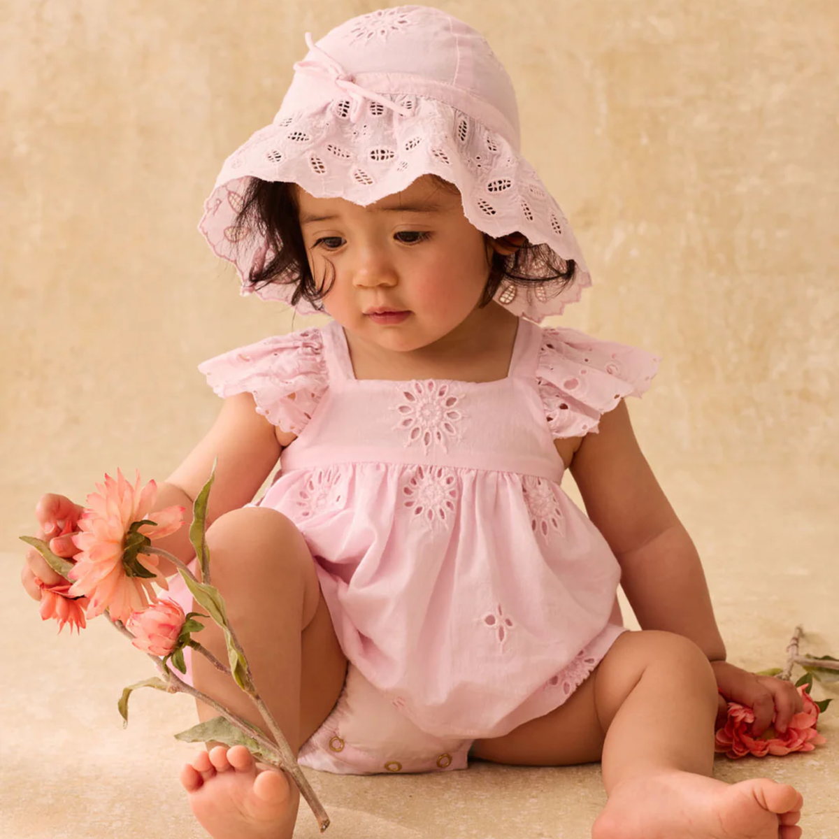 Baby in a pink lacey outfit and hat holding flowers on a beige background.