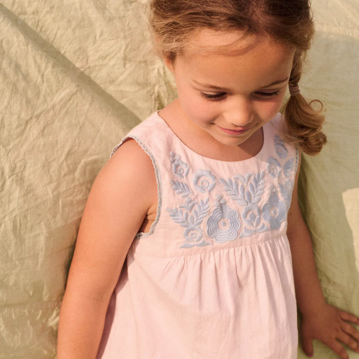Young girl wearing a light pink top with blue embroidery against a textured beige wall.