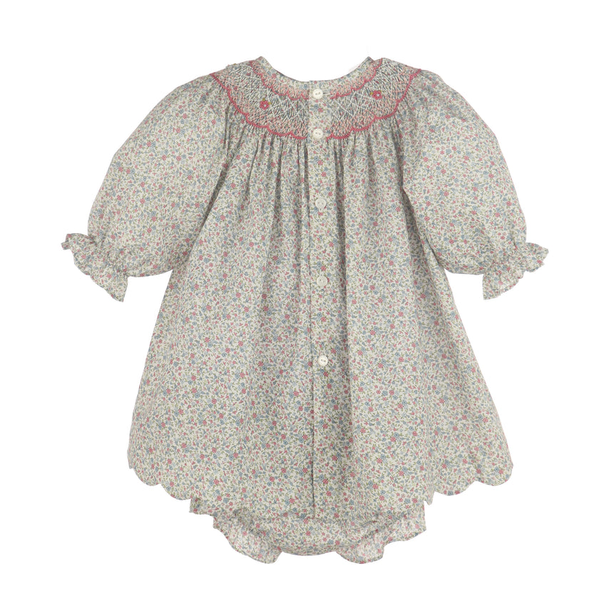 A girl's smocked dress with short puff sleeves, a scalloped hem, and a tiny floral print on a light gray background with pink and green details. It features a smocked neckline with pink embroidered trim and decorative buttons down the back.