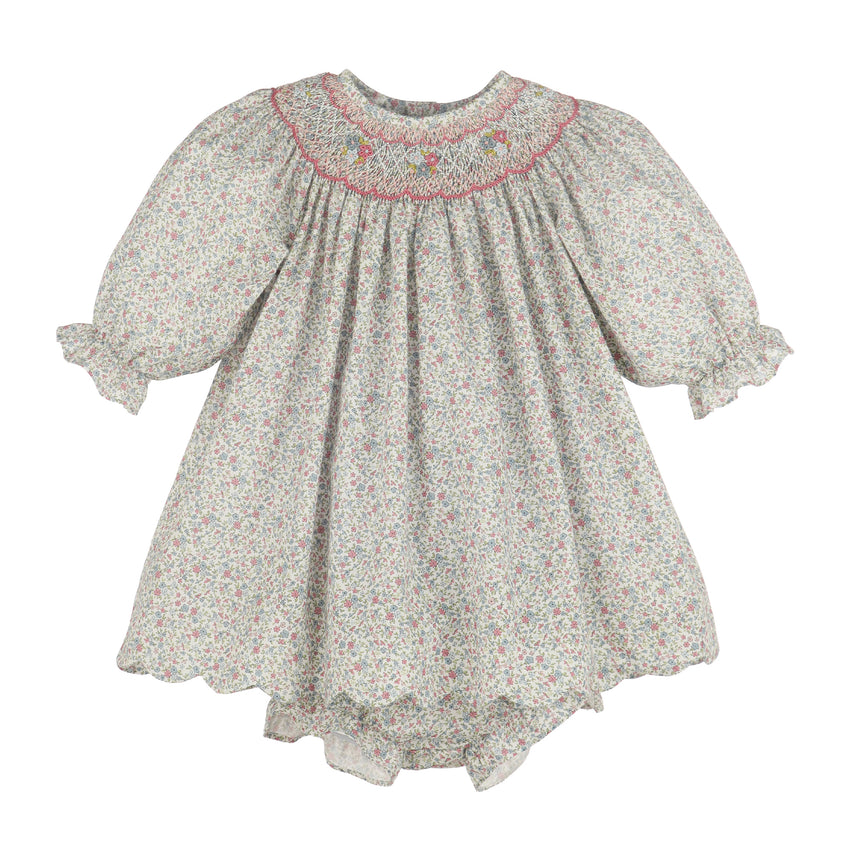 A girl's smocked dress featuring a delicate multicolor floral print on a light grayish-beige background, with a pink embroidered neckline and ruffled sleeves.