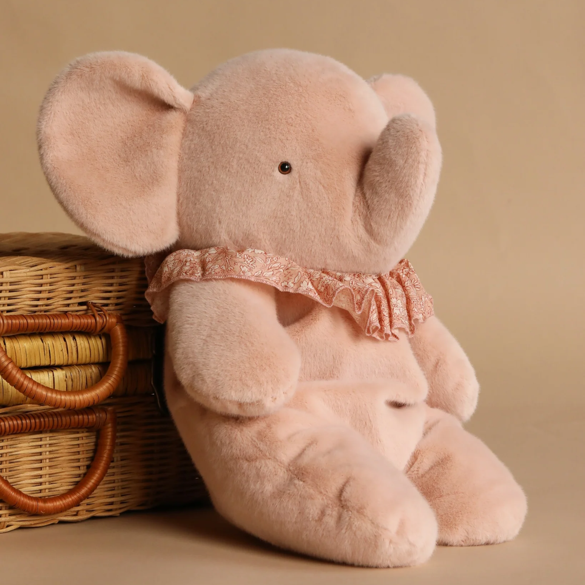Plush elephant toy with a lace collar next to a wicker basket on a beige background.