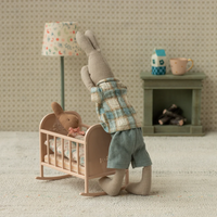 Stuffed animal in a plaid shirt and shorts standing next to a baby crib with another stuffed animal inside, in a room setting.
