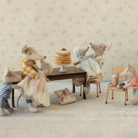 Stuffed animal mice gathered around a table with a cake in a room with floral wallpaper.