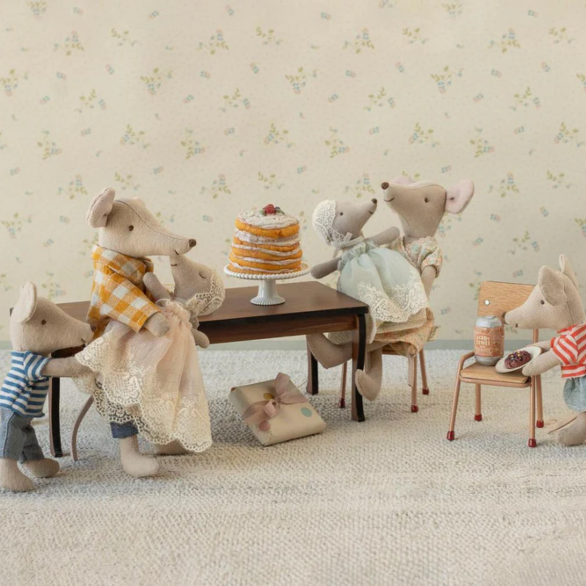 Stuffed animal mice gathered around a table with a cake in a room with floral wallpaper.