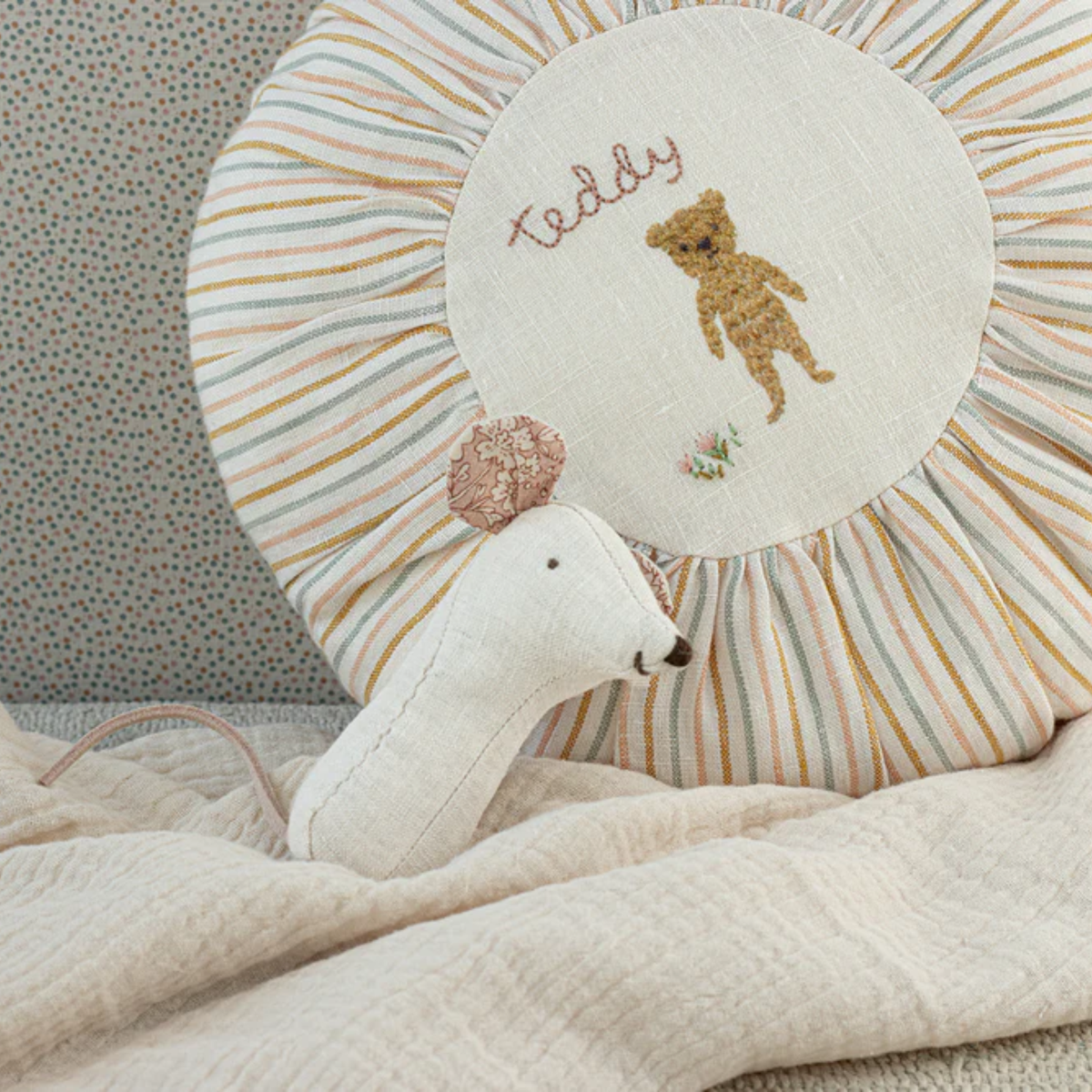 White mouse rattle on a bed with a "teddy" embroidered pillow and resting on a blanket.