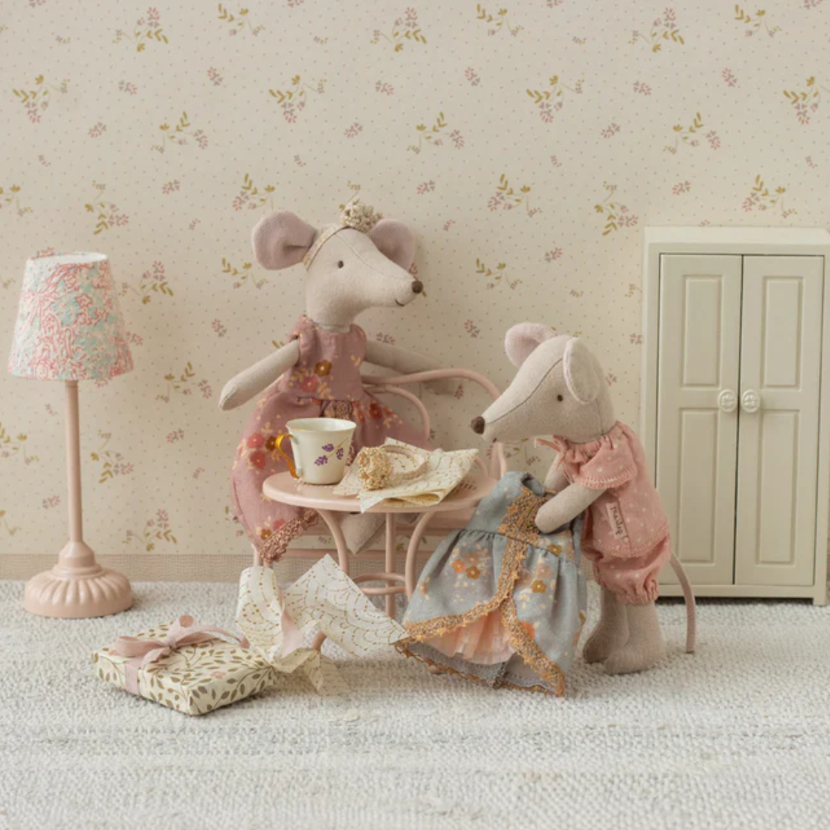 Two plush mice in a decorative room with floral wallpaper, a lamp, table and opening a dress gift.