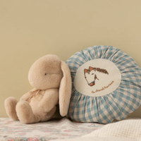 Plush toy bunny next to a checkered pillow with an embroidered horse on a soft surface.