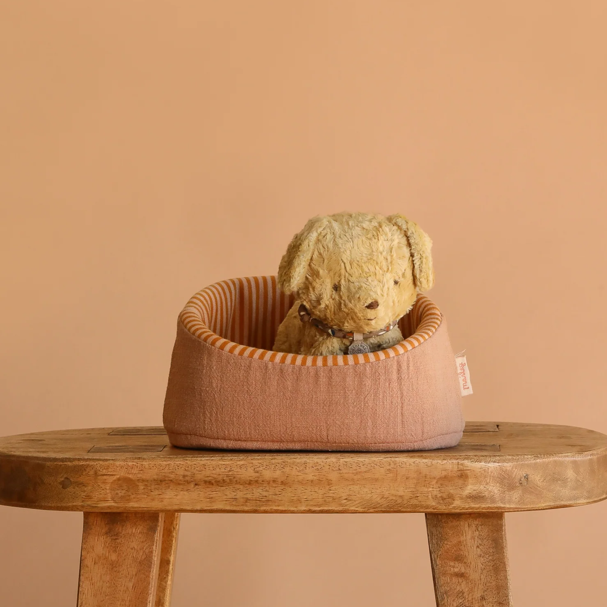 Toy stuffed brown dog in a pink pet bed on a wooden stool against a beige background.