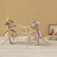Two toy mice in ballet costumes standing next to a small white bench against a beige wall.