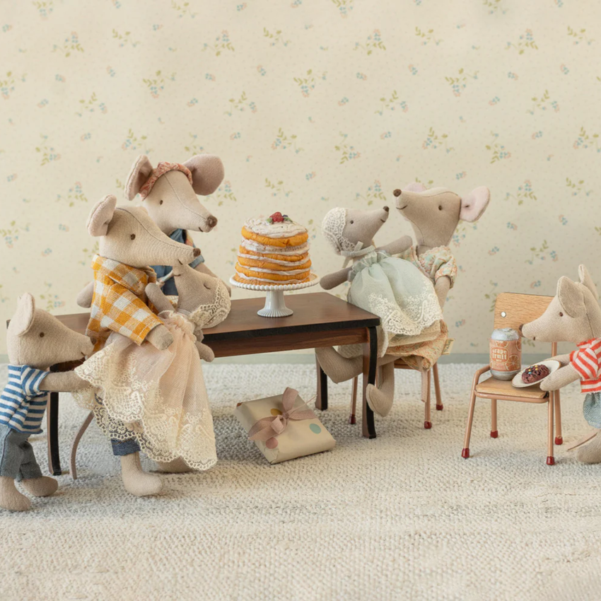 Stuffed toy mice family gathered around a table with a cake, set against a floral wallpaper background.