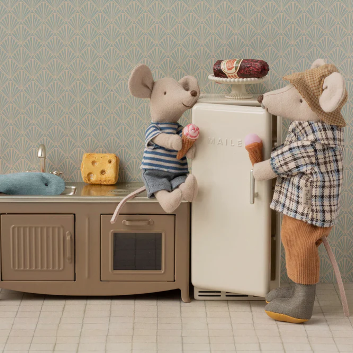 Two toy mice in a kitchen setting with a refrigerator, cheese, ice cream  and sink.