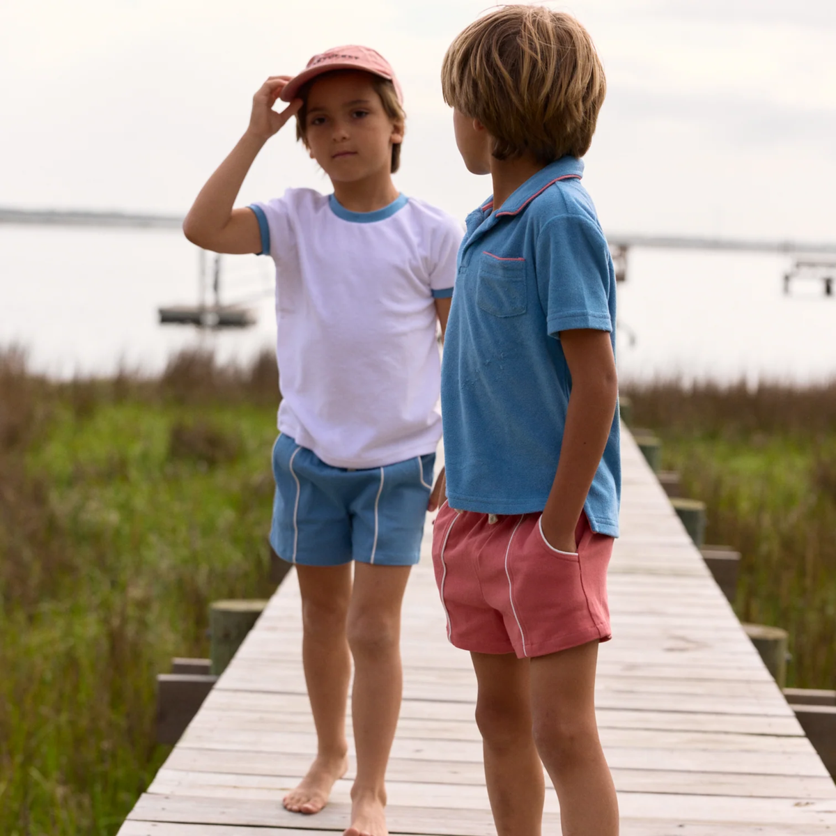 Two children standing on a wooden dock One in white t-shirt with blue trim and blue shorts with white trim. Other in blue t-shirt, red trim and red shorts with white trim.