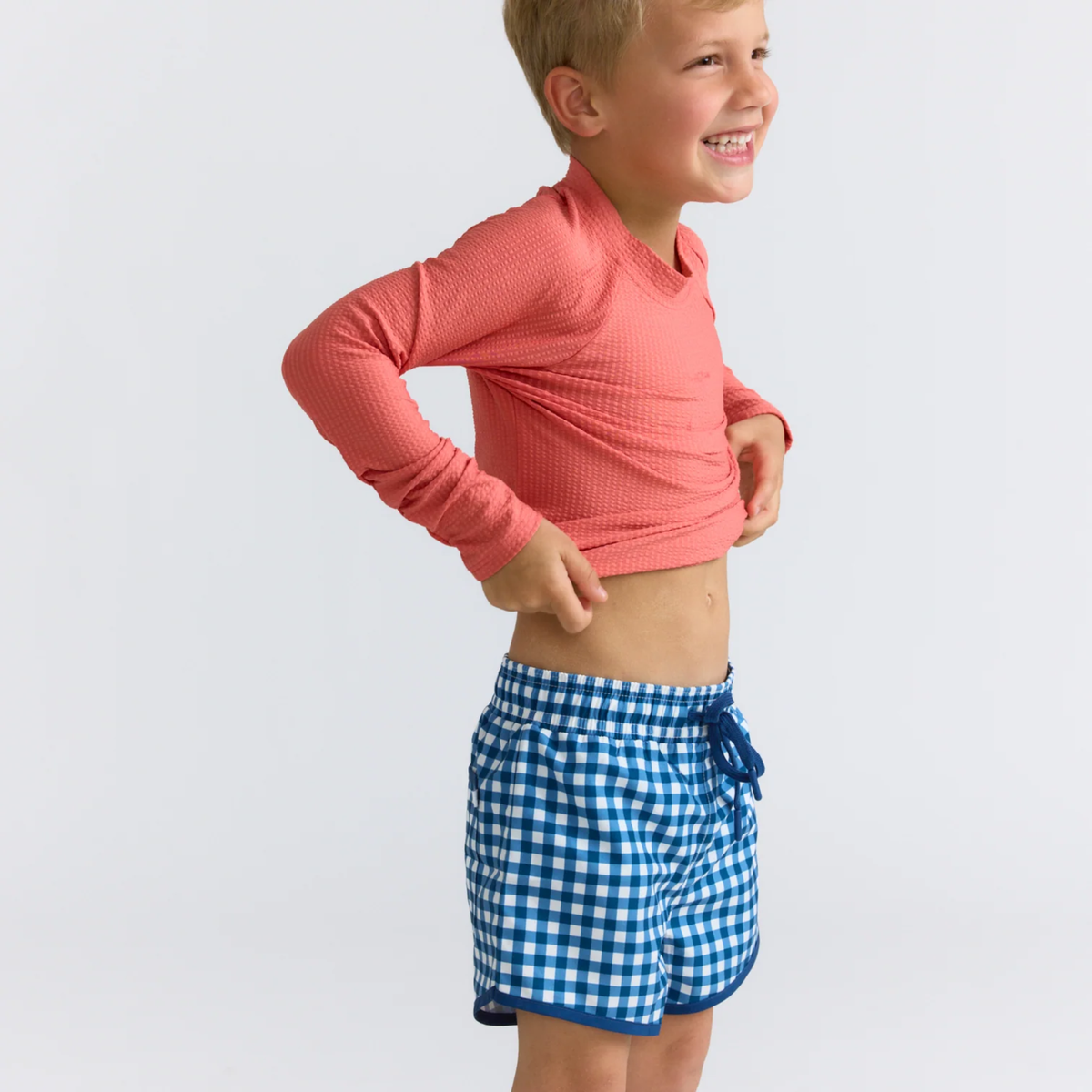 Child wearing a pink long-sleeve shirt and blue checkered boardie shorts on a light gray background