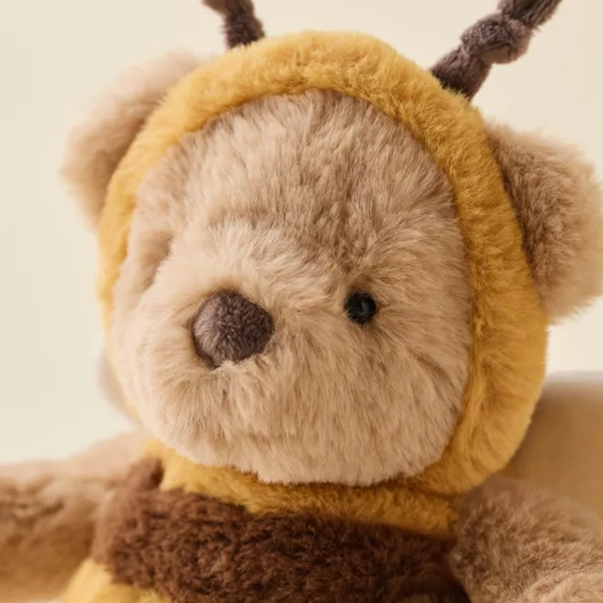 A close-up of a plush teddy bear wearing a yellow bee hood with brown antennae and a brown scarf, set against a soft cream background.