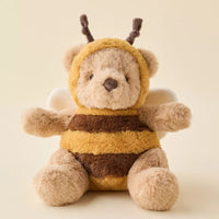 A plush teddy bear dressed as a bee, featuring a soft beige body, yellow and brown striped fuzzy suit, and small white wings, photographed against a warm cream background.