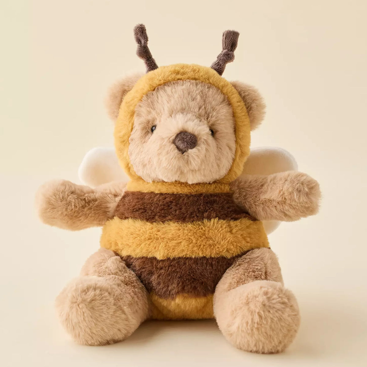 A plush teddy bear dressed as a bee, featuring a soft beige body, yellow and brown striped fuzzy suit, and small white wings, photographed against a warm cream background.