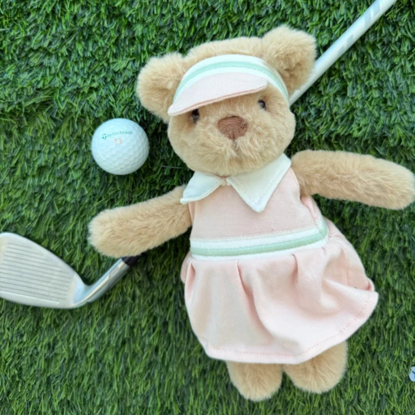 Teddy bear in a pink golf dress with a white collar and visor on a grass background.