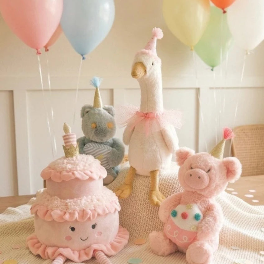 A soft plush toy set arranged on a knitted blanket with pastel balloons in the background. The set includes a pink tiered cake plush with a candle, a white goose wearing a pink tutu, a gray bear wearing a party hat, and a pink pig plush in a pink outfit.
