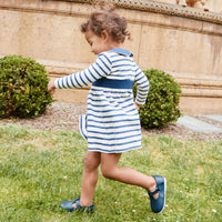 A toddler girl wearing a blue and white striped long-sleeve dress and navy blue shoes is walking on a grassy lawn in front of a stone wall.
