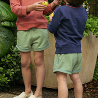 Two children outdoors wearing light sage green shorts with contrasting blue piping, paired with long-sleeve sweatshirts in coral pink and navy blue.