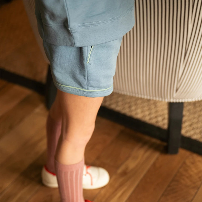 A child wearing light blue shorts and a matching top, paired with pink ribbed socks and white sneakers, standing on a wooden floor next to a striped upholstered chair.