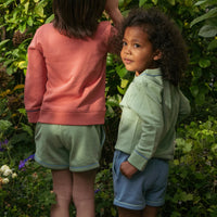 Two children outdoors in a lush green garden, wearing matching casual outfits. One child is shown from the back in a coral-pink sweatshirt and sage green shorts, while the other child looks over their shoulder wearing a sage green zip-up jacket and blue shorts.