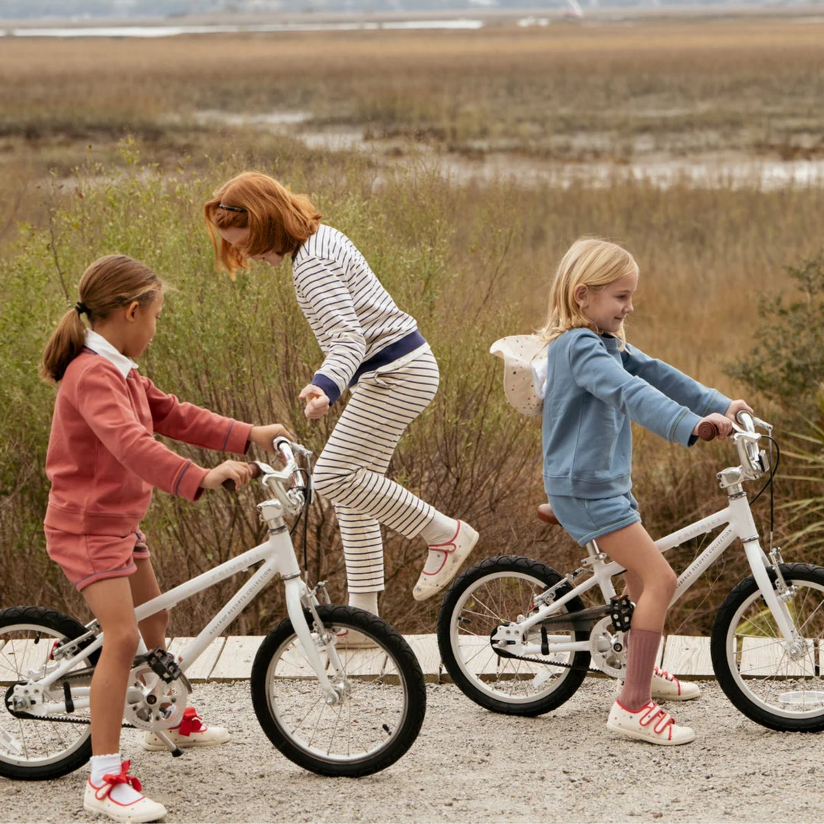 Three children riding or standing beside white bicycles on a gravel path in an outdoor marshland setting. The children wear coordinated pastel and striped outfits.