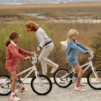 Three children riding or standing beside white bicycles on a gravel path in an outdoor marshland setting. The children wear coordinated pastel and striped outfits.