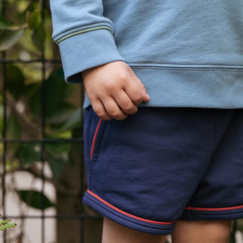 A close-up of a child wearing navy blue shorts with red piping trim and a light blue sweatshirt, standing outdoors in front of a green wire fence.