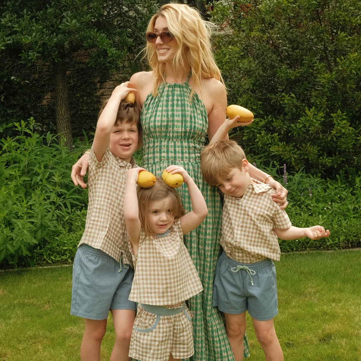 A family of four posing outdoors in a grassy garden. The mother wears a green and white checkered halter maxi dress, while the three children wear matching beige and white gingham check tops and shorts in blue denim.