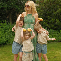 A family of four posing outdoors in a grassy garden. The mother wears a green and white checkered halter maxi dress, while the three children wear matching beige and white gingham check tops and shorts in blue denim.