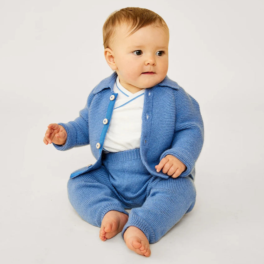 A baby sitting and wearing a blue knit cardigan and matching knit pants over a white top, photographed against a plain light background.