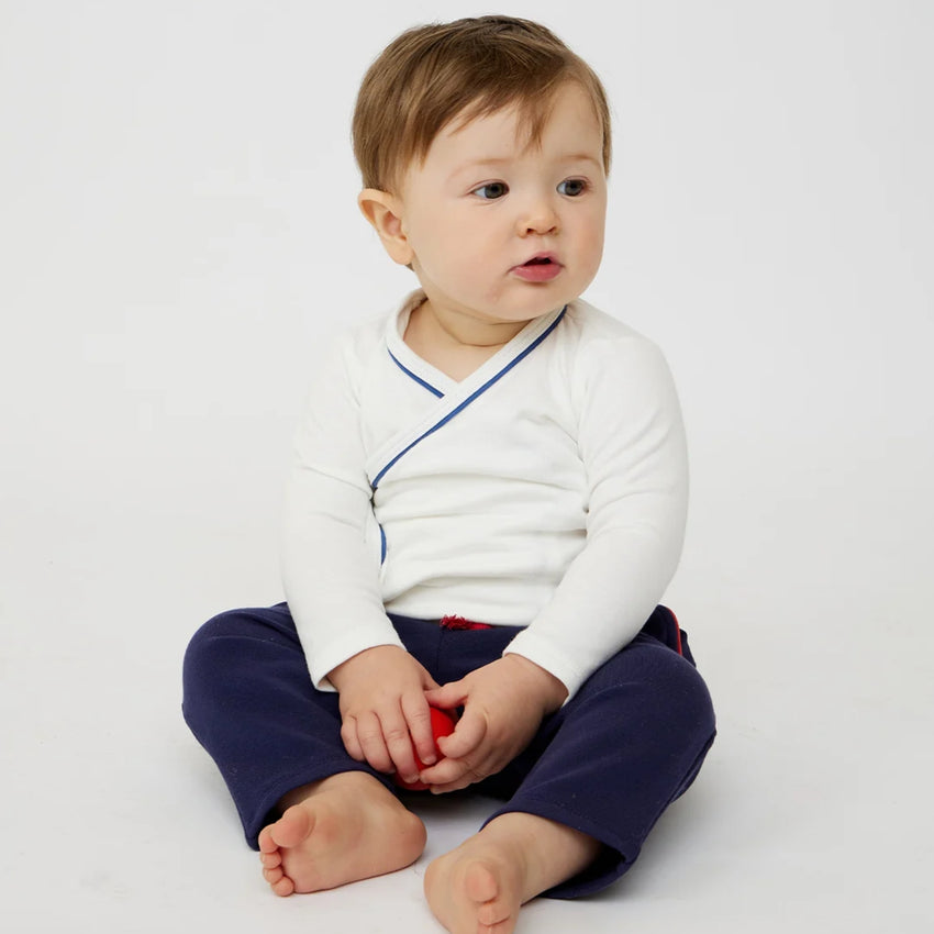 A baby sitting cross-legged on a white studio background, wearing a white long-sleeve crossbody wrap-style top with thin blue piping and navy pants.