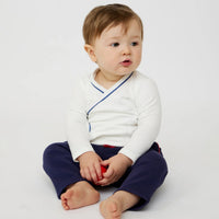 A baby sitting cross-legged on a white studio background, wearing a white long-sleeve crossbody wrap-style top with thin blue piping and navy pants.