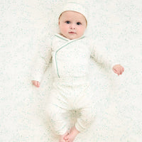 A baby lying on their back wearing a long-sleeve crossbody wrap-style romper and matching hat, both in an off-white color with a light green star pattern and green piping, on a matching speckled blanket.