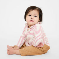 A baby sitting cross-legged on a white studio background, wearing a pink and white striped long-sleeve top with a ruffled collar and tan pants.