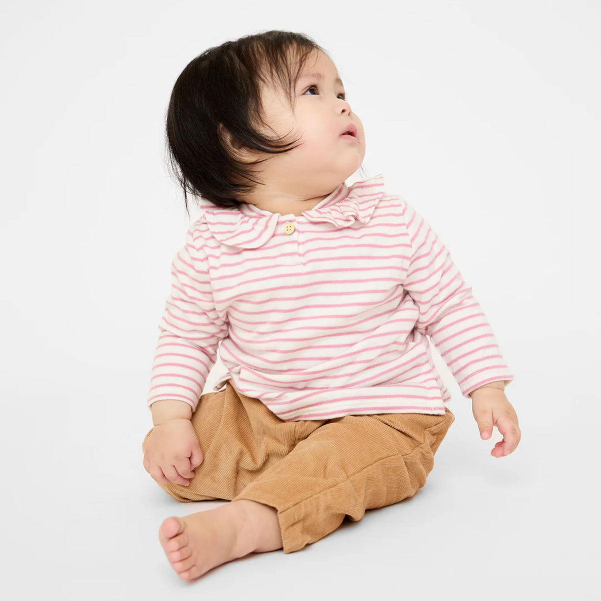 A baby sitting on a white studio background, wearing a pink and white striped long-sleeve top with a ruffled collar and tan pants.