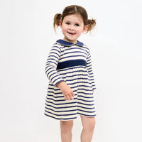 A young girl with brown hair in pigtails smiling while wearing a long-sleeve navy and cream striped dress with a solid navy collar, set against a plain white studio background.