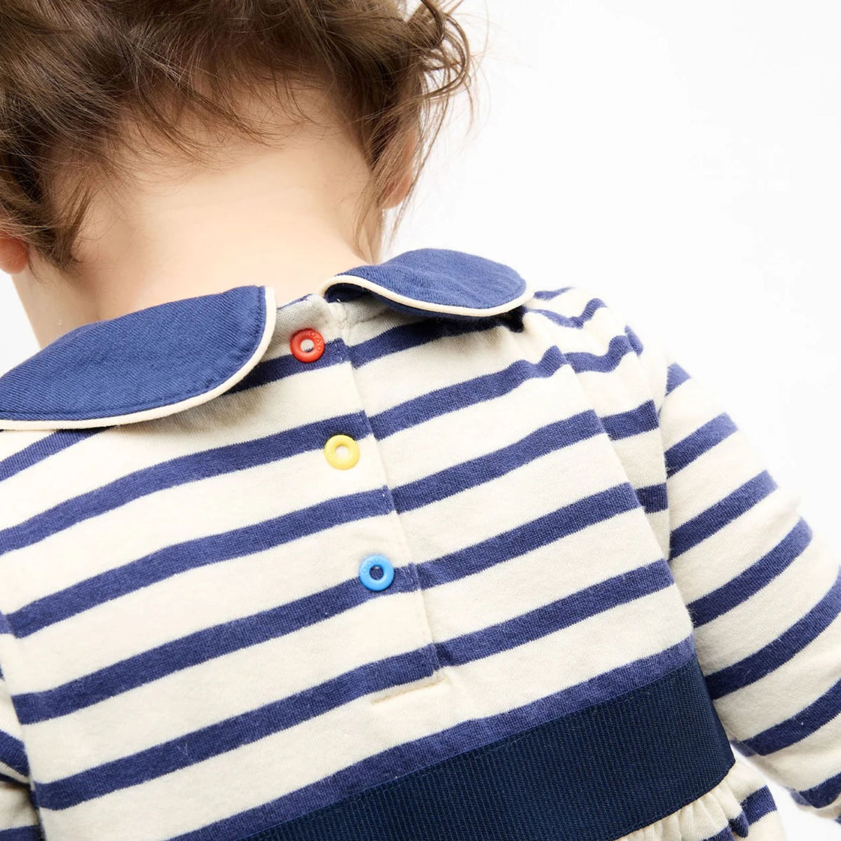 Close-up back view of a child wearing a cream and navy blue horizontally striped long-sleeve top with a blue collar and colorful snap buttons.
