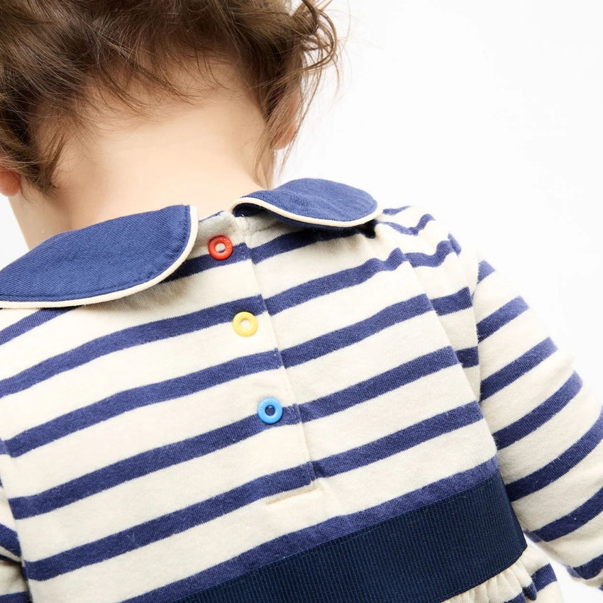 Close-up back view of a child wearing a cream and navy blue horizontally striped long-sleeve top with a blue collar and colorful snap buttons.